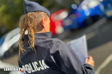 Policjantka w mundurze z napisem „Policja” stoi tyłem na drodze, trzymając dokument, w tle widać policyjny radiowóz.